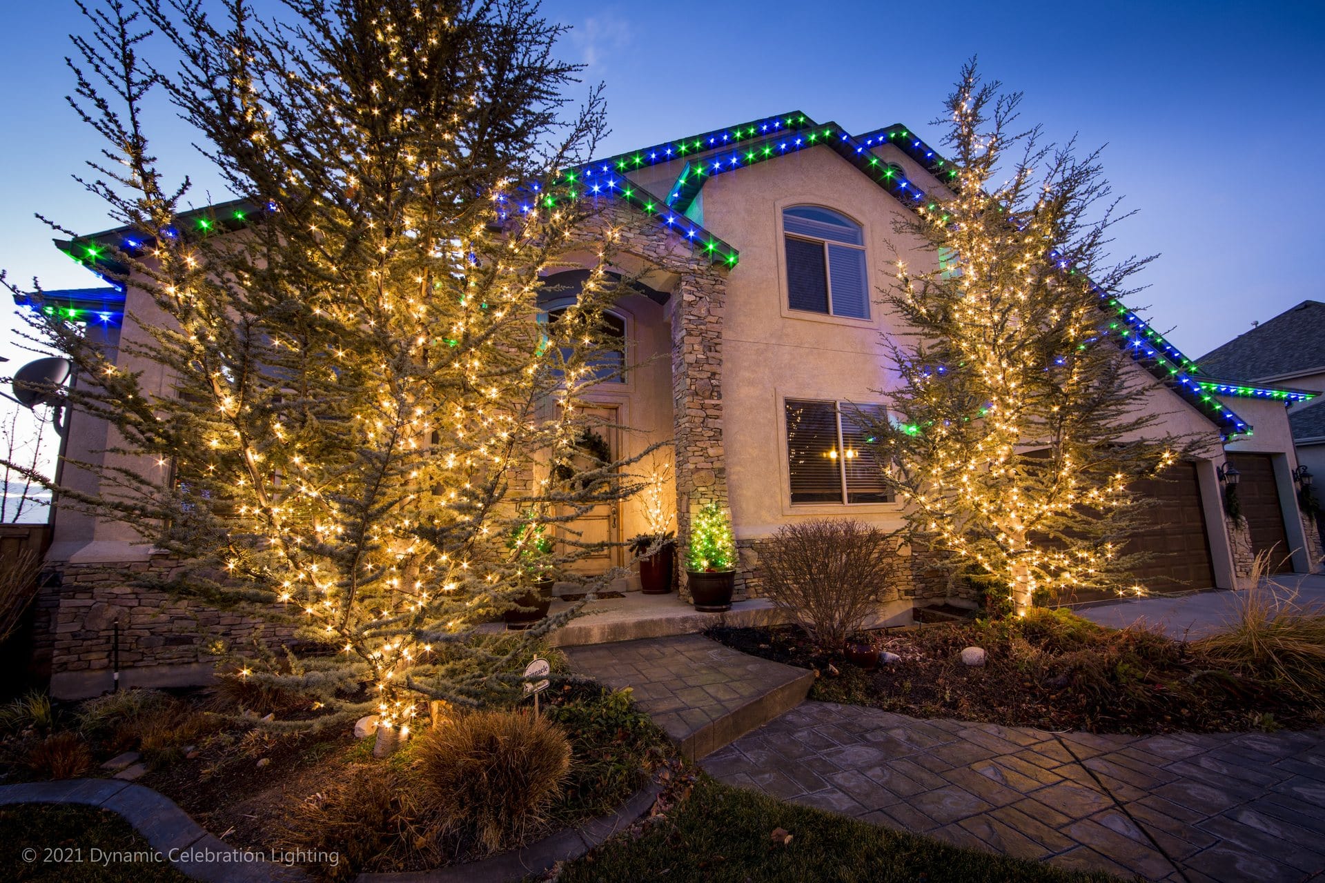 A house with two lit trees and colorful LEDs along the roof, taken at dusk.