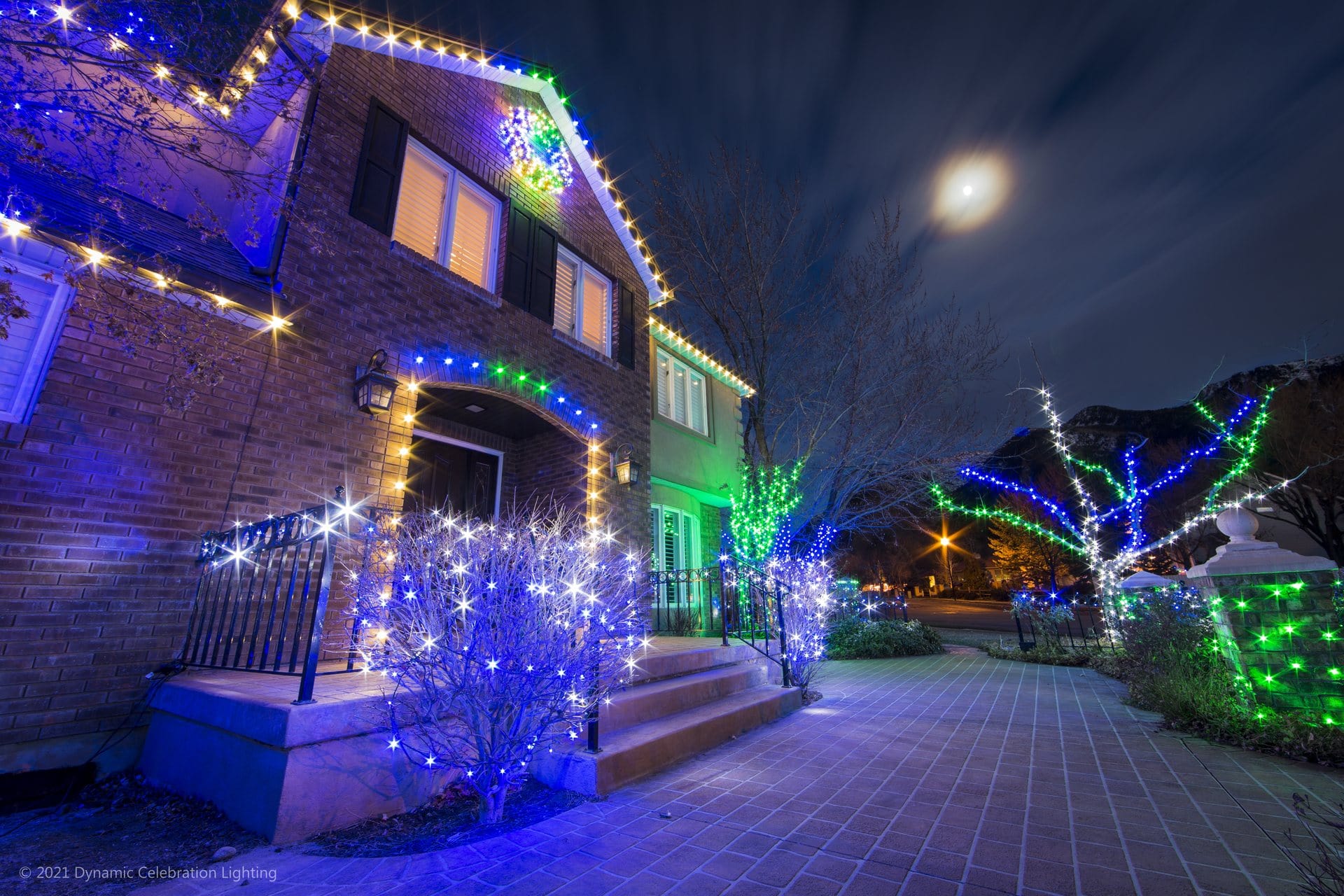 A house and trees decorated with blue, green, and white holiday lights under a cloudy night sky.