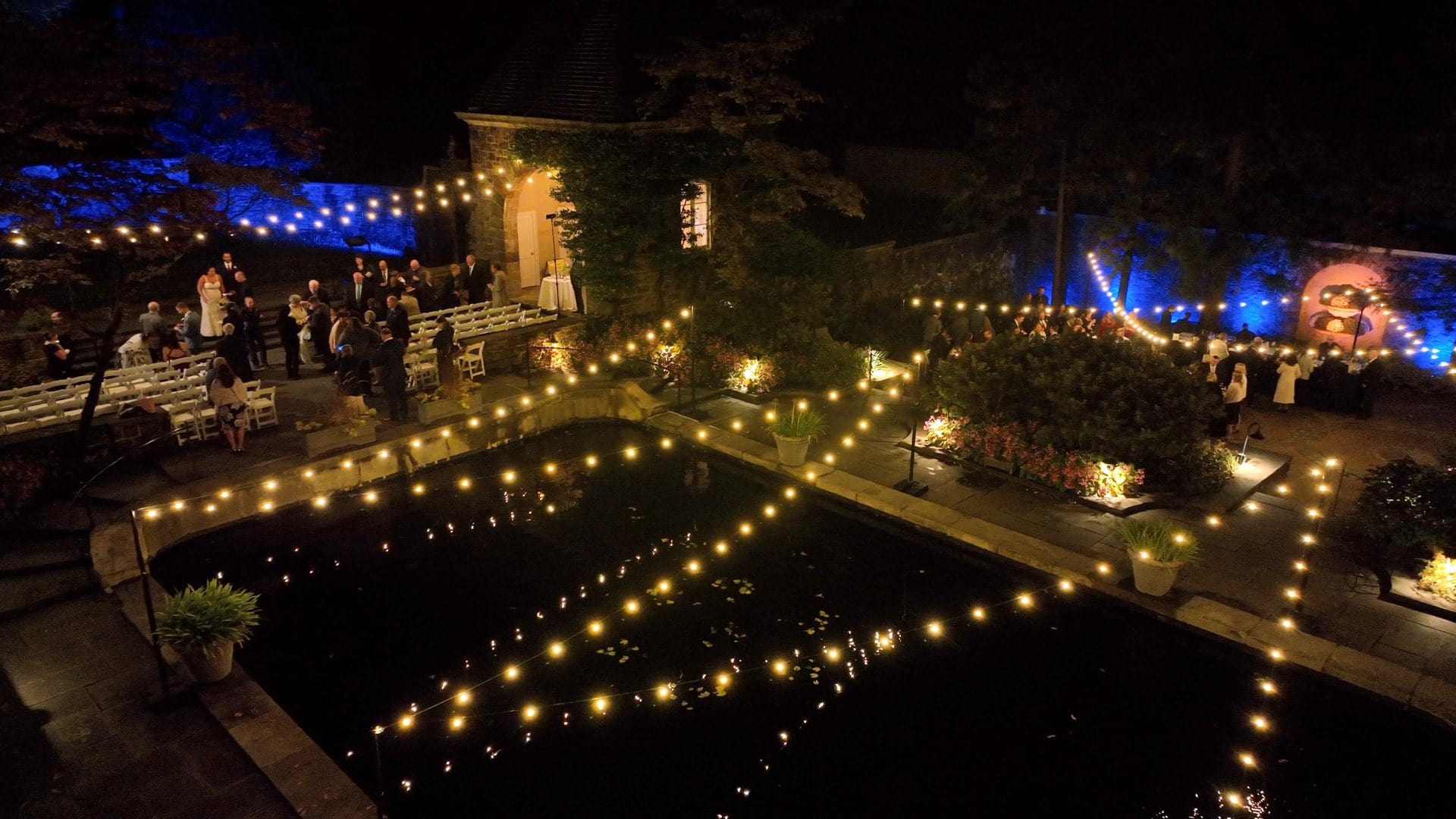 Nighttime outdoor wedding with string lights over a pond and guests gathered near trees and a stone building.