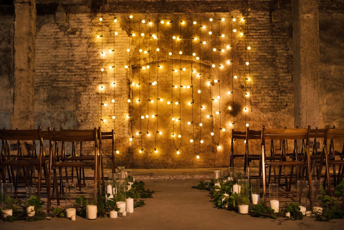 Wooden chairs face a wall with string lights; greenery and candles line the aisle in a rustic setting.