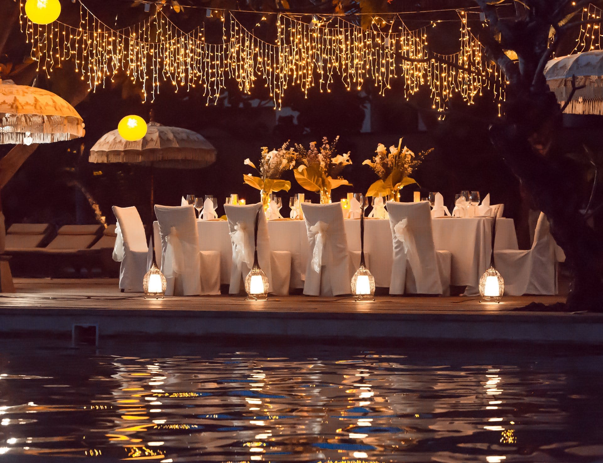 Elegant outdoor dinner setup with white tablecloths, floral centerpieces, and string lights by the water.