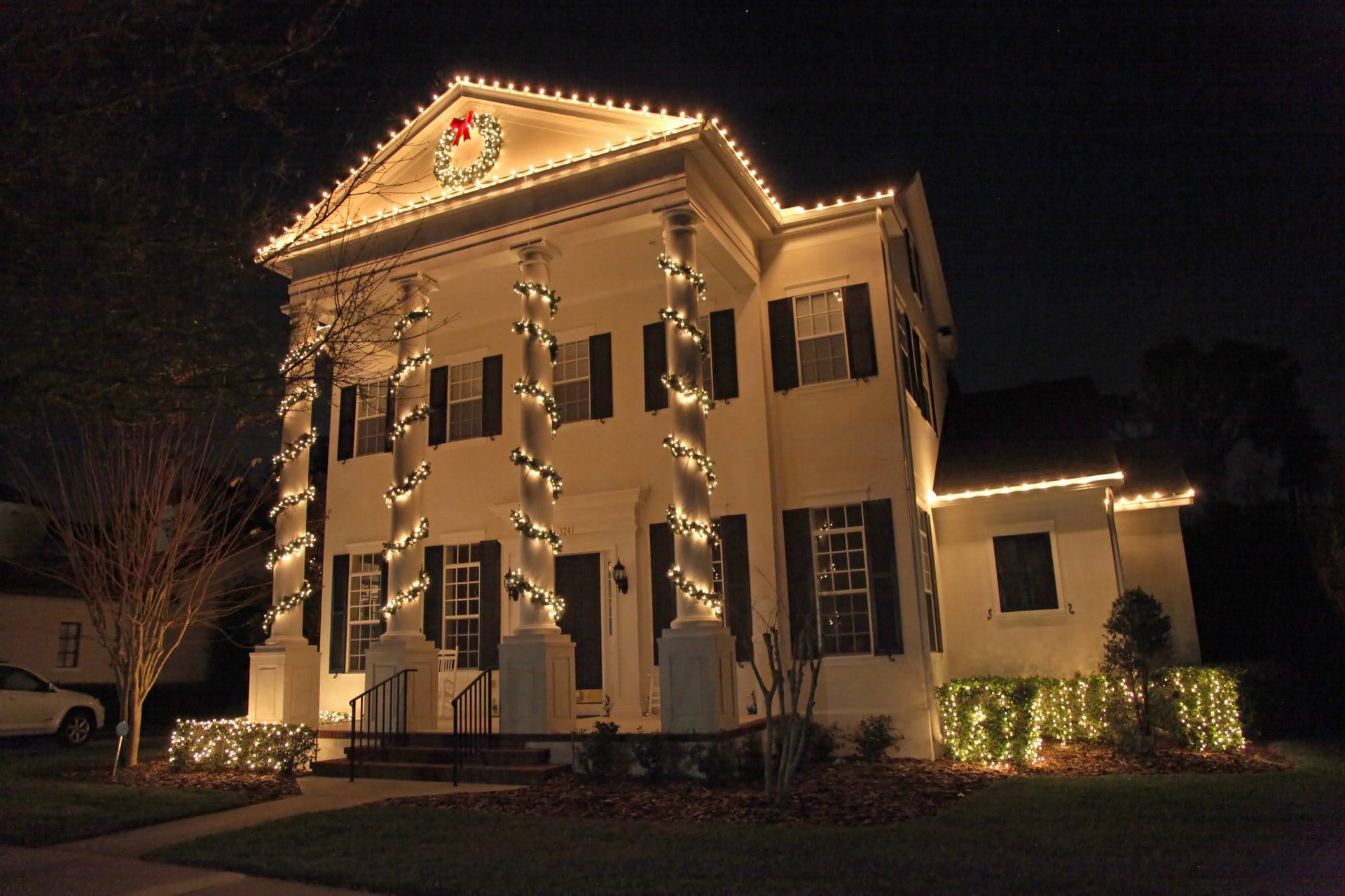 Two-story house at night with Christmas lights, wreath, and garlands on columns and roofline.