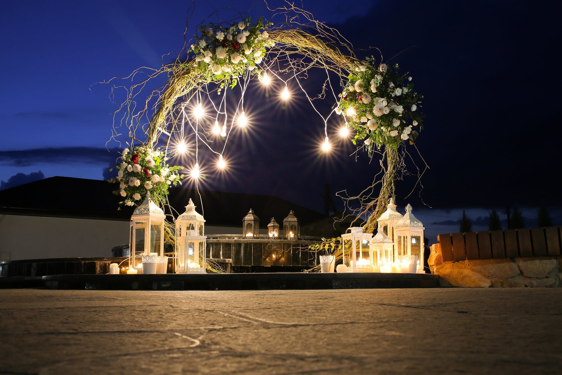 A floral arch with hanging lights and lanterns is set up outdoors at dusk, creating a romantic scene.