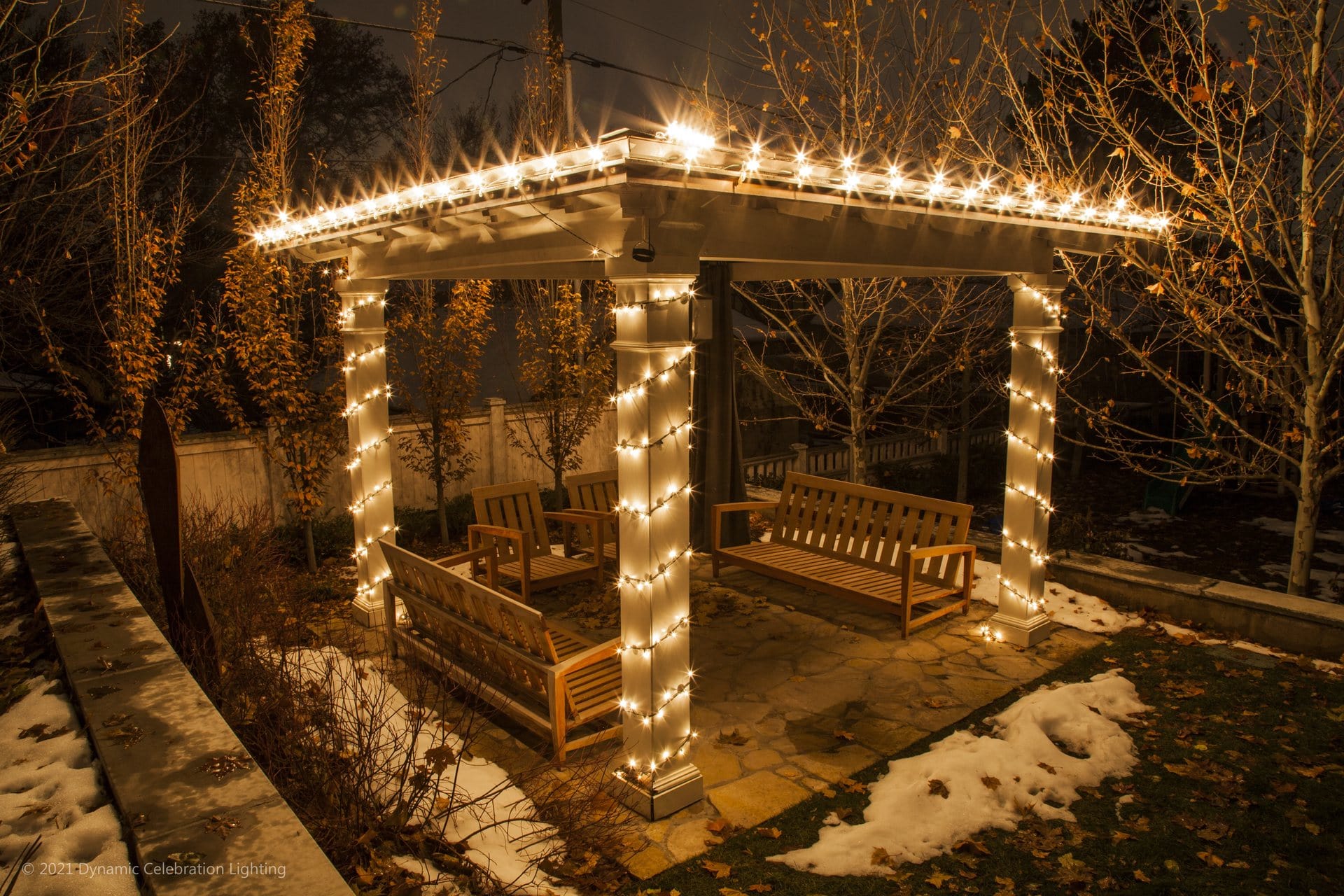 A wooden gazebo with benches is wrapped in string lights at night, surrounded by patches of snow and trees.