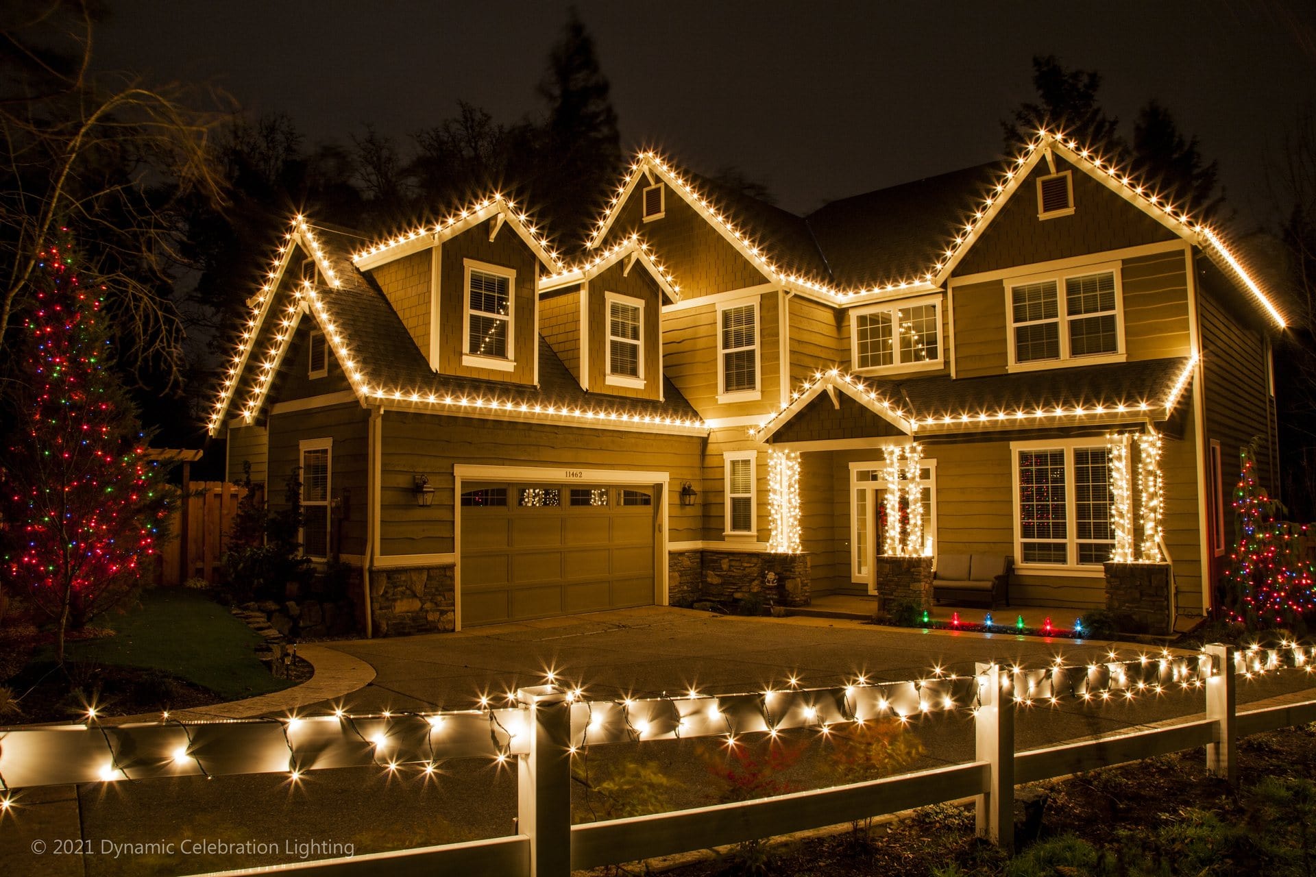 A two-story house decorated with white Christmas lights outlining the roof, windows, and fence at night.