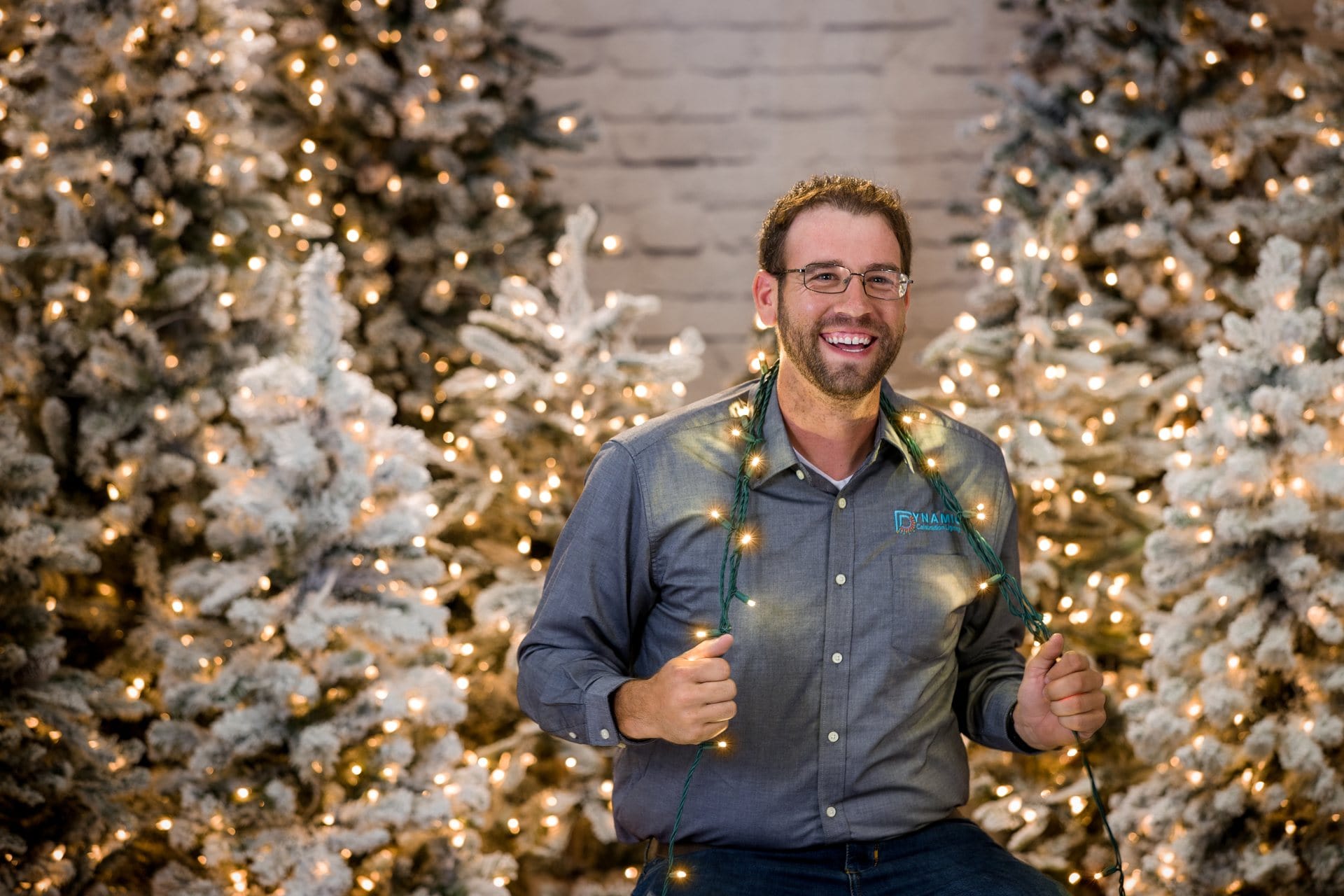 Smiling man with glasses holds string lights, sitting between snow-covered, decorated Christmas trees.