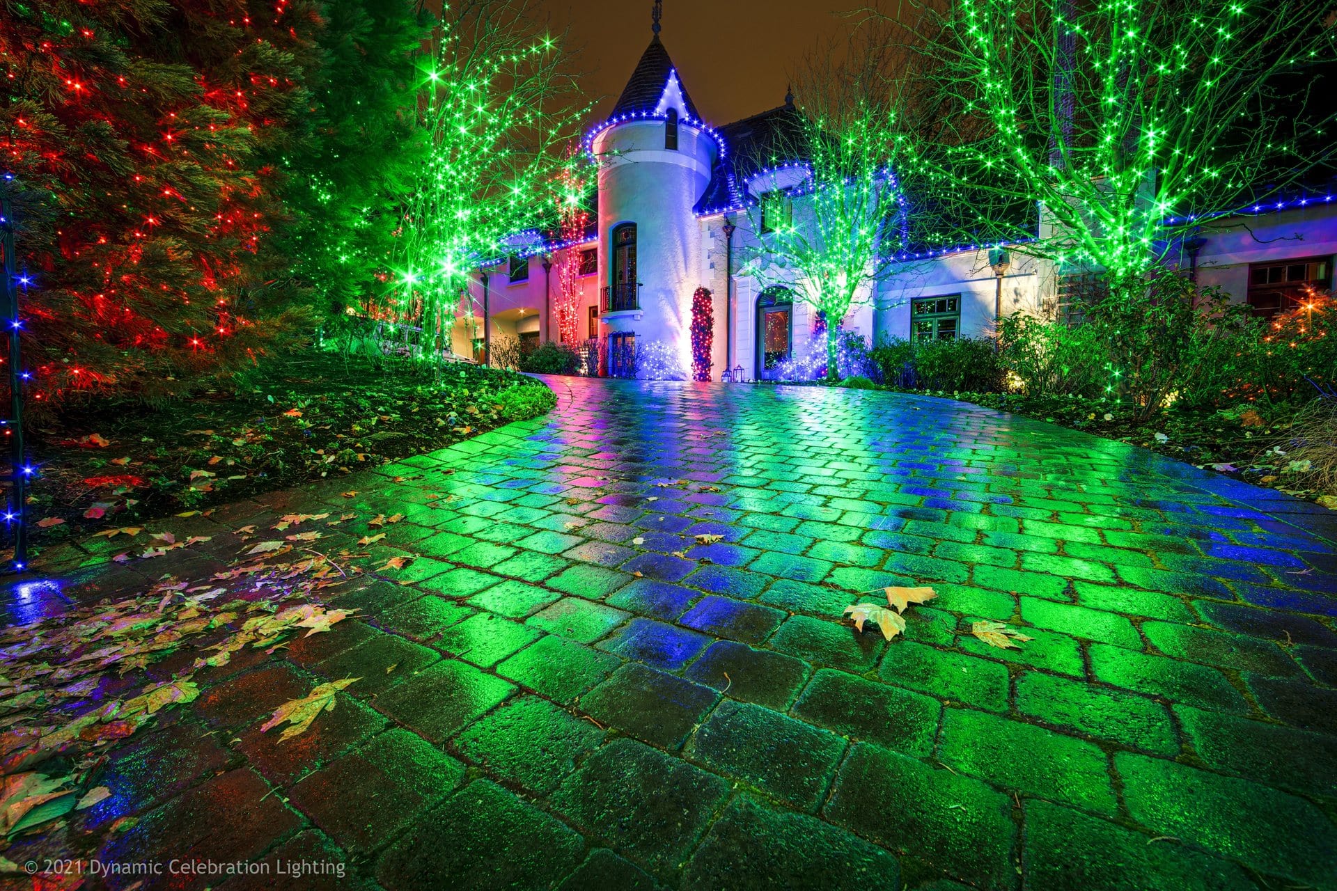 A house and driveway lit up with colorful holiday lights at night, reflecting on wet cobblestones.