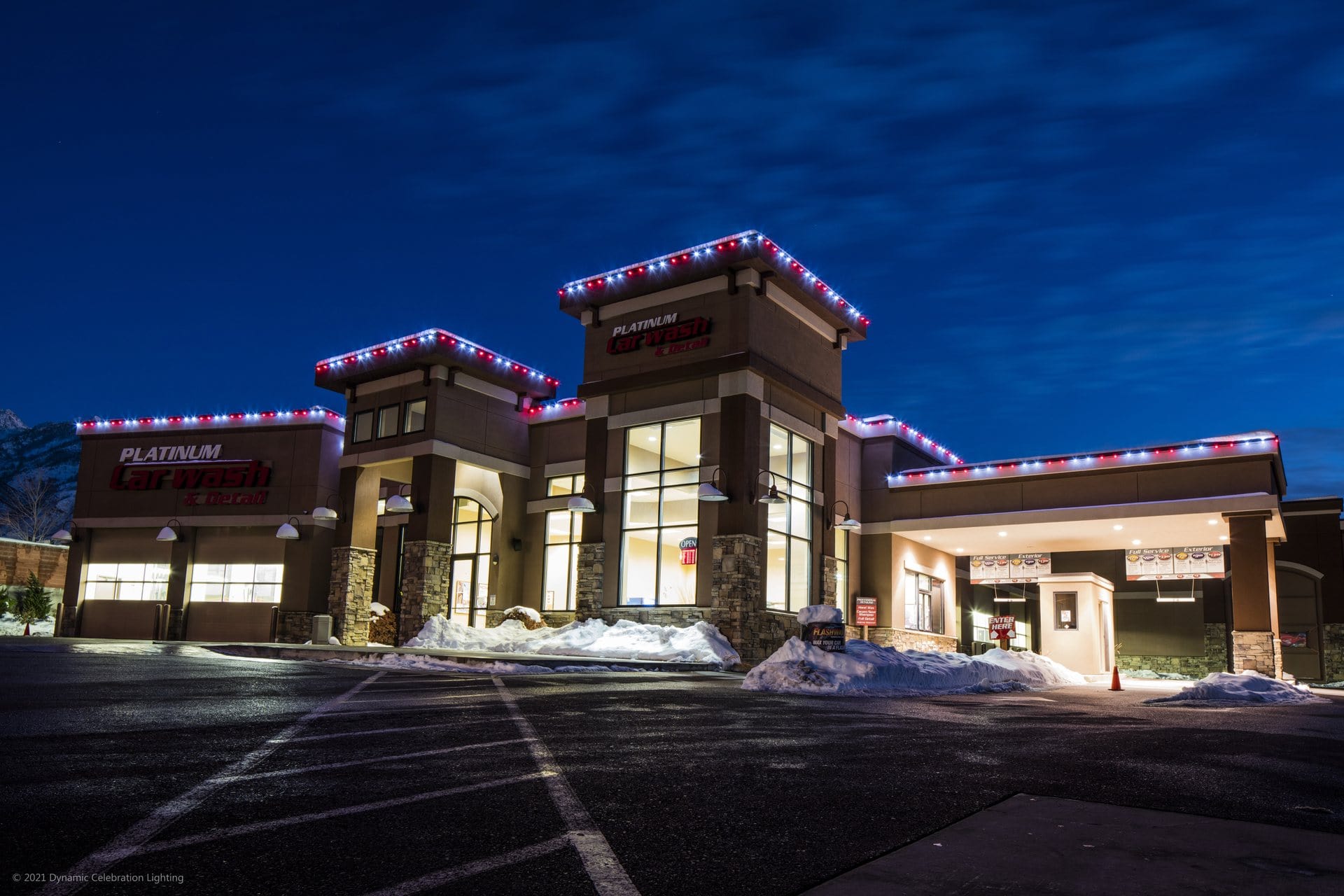 A modern car wash building at night, lit with red and white lights, snow piled around the parking lot.