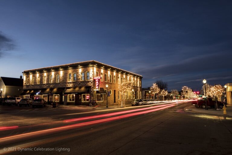 A café in a brick building with holiday lights at dusk; car light trails on the street in front.