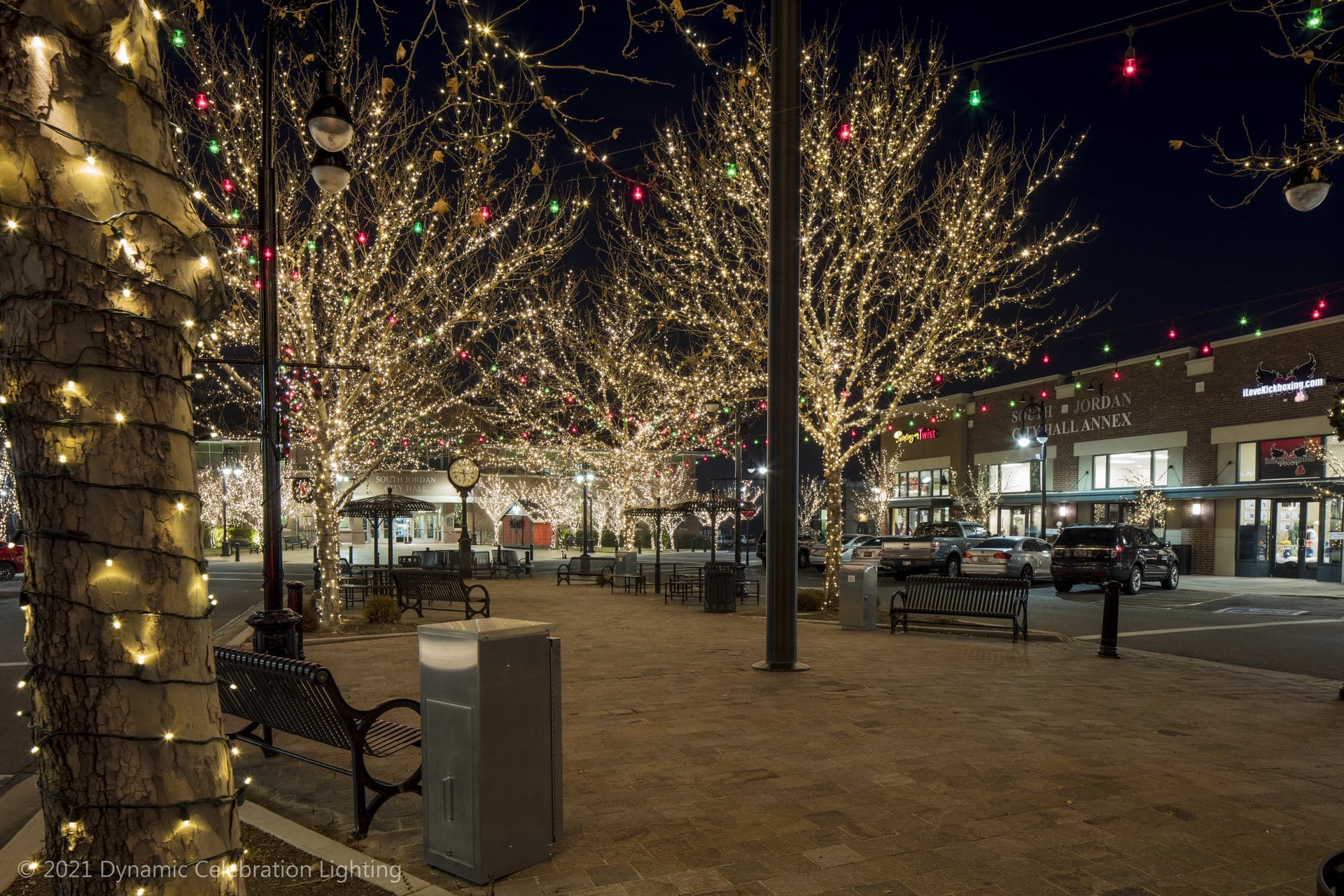 A town square at night with trees and buildings decorated with festive string lights.