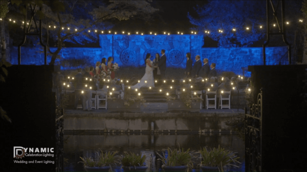 A wedding ceremony at night with string lights, blue lighting, and guests seated outdoors by a pond.