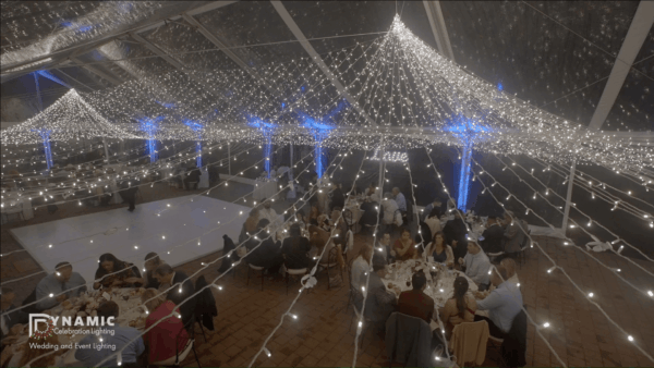 Guests seated at round tables under a canopy of string lights at an indoor event or wedding reception.