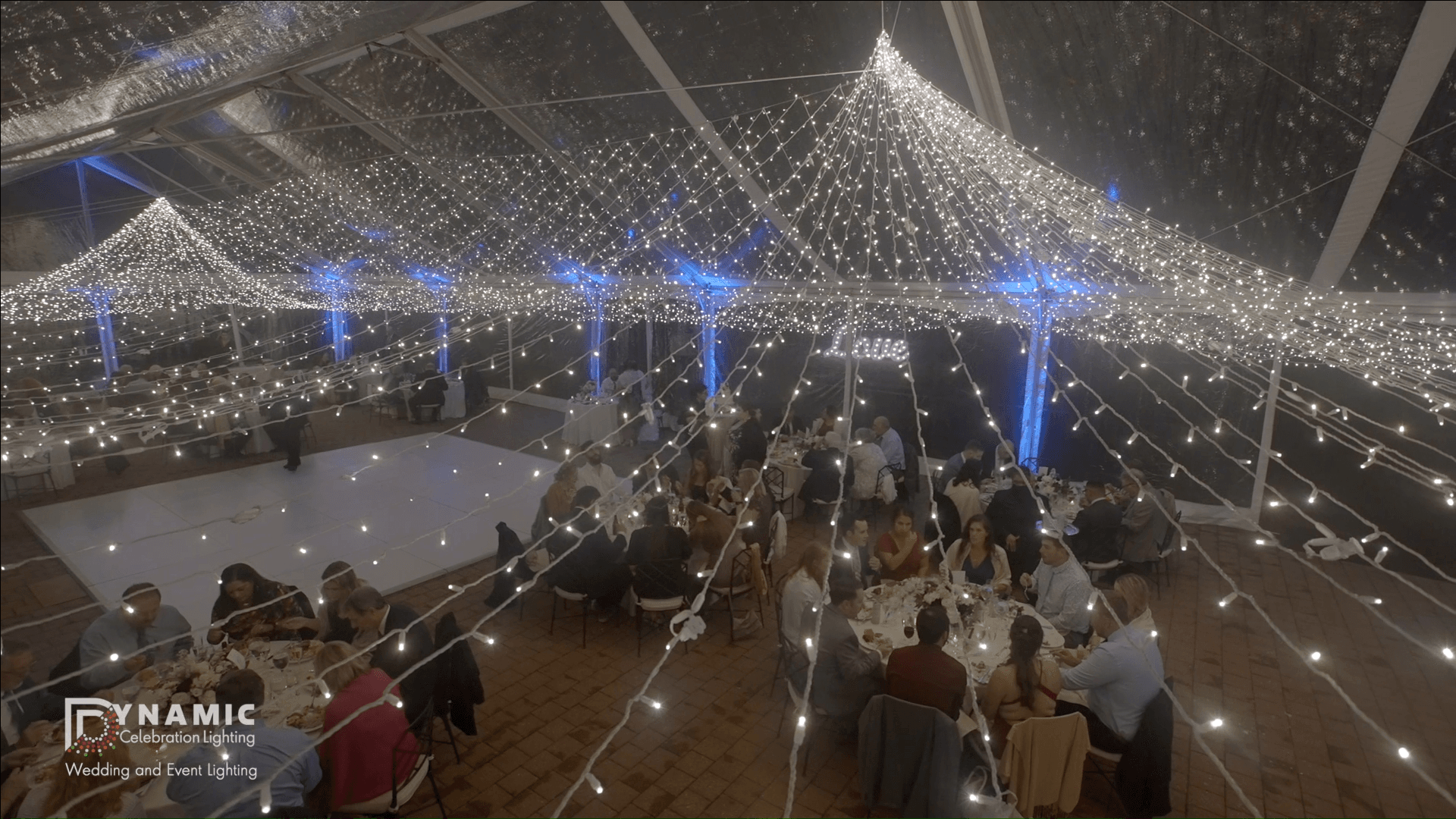 Guests seated at round tables under a canopy of string lights at an indoor event or wedding reception.