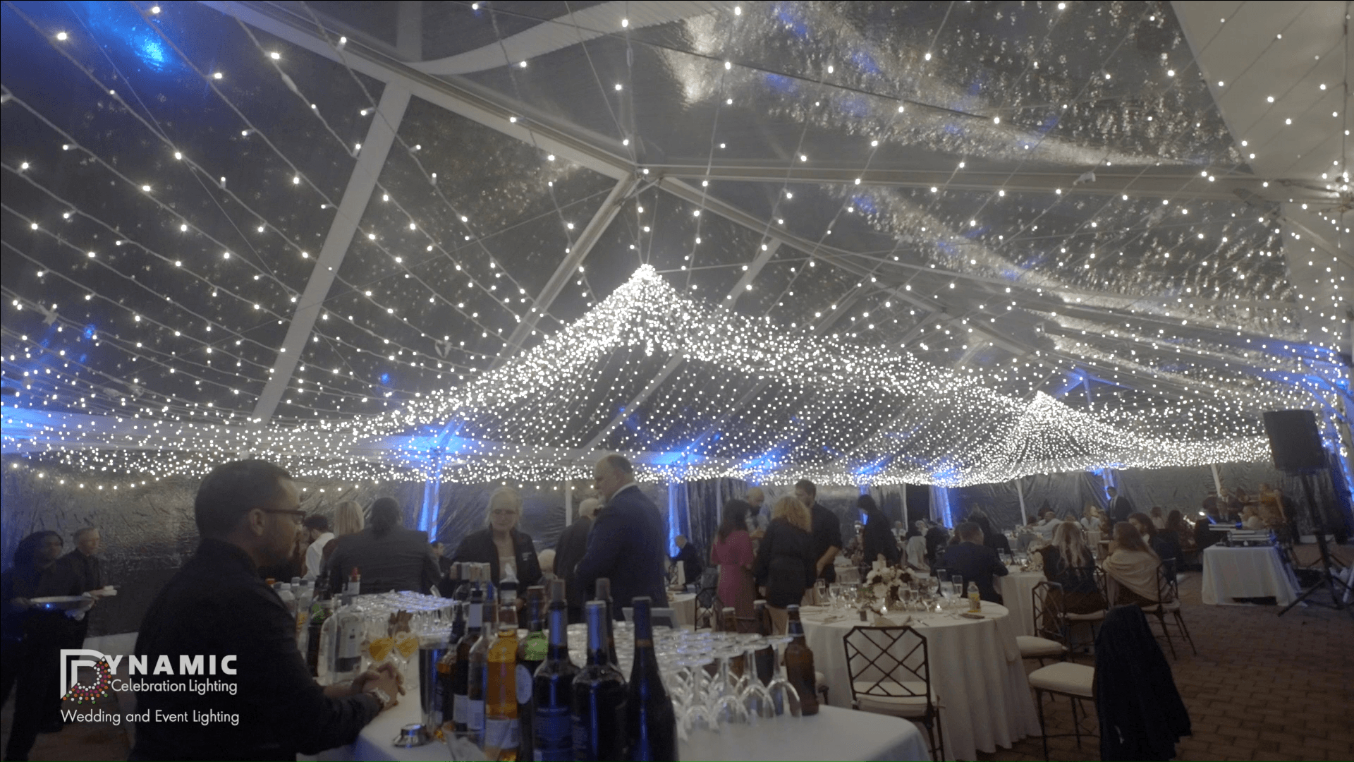 People at an elegant event under a tent with string lights and tables set with drinks and glasses.