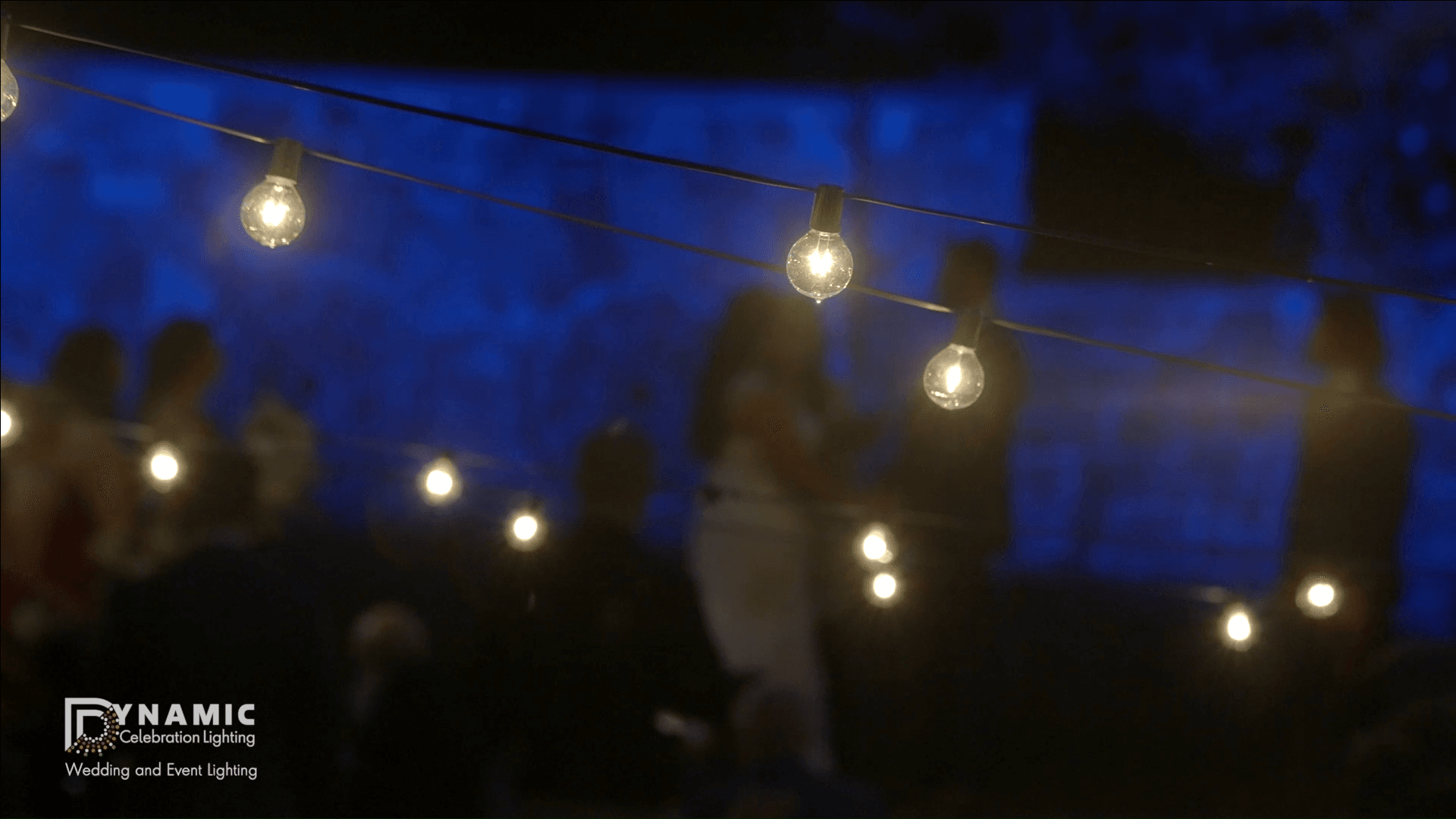 String lights in focus with blurry figures and blue-lit background at an outdoor evening event.