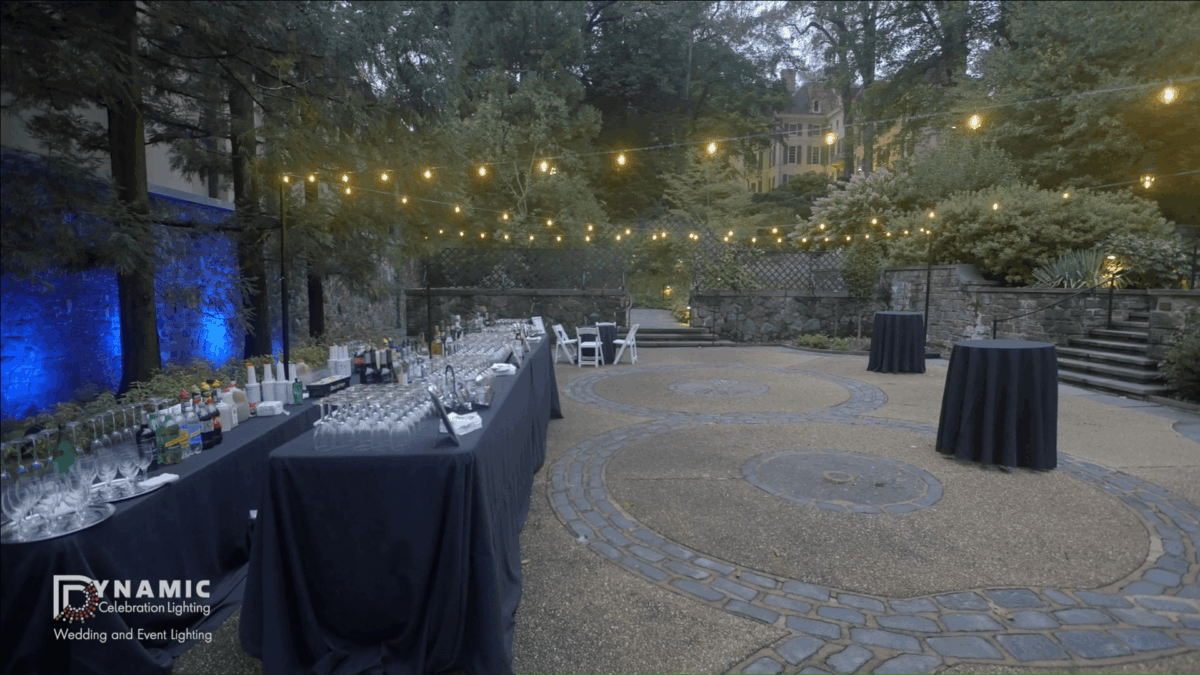 Outdoor event setup with string lights, cocktail tables, and a bar under trees in the evening.