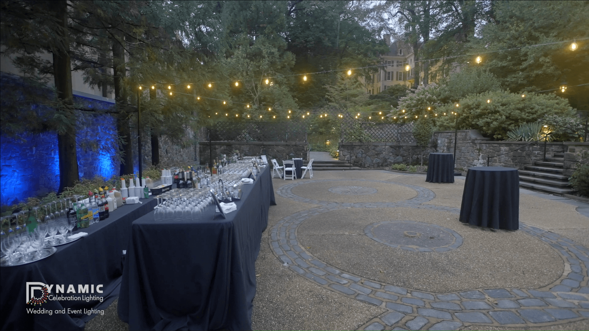 Outdoor event space at dusk with string lights, cocktail tables, and drink stations set up for guests.