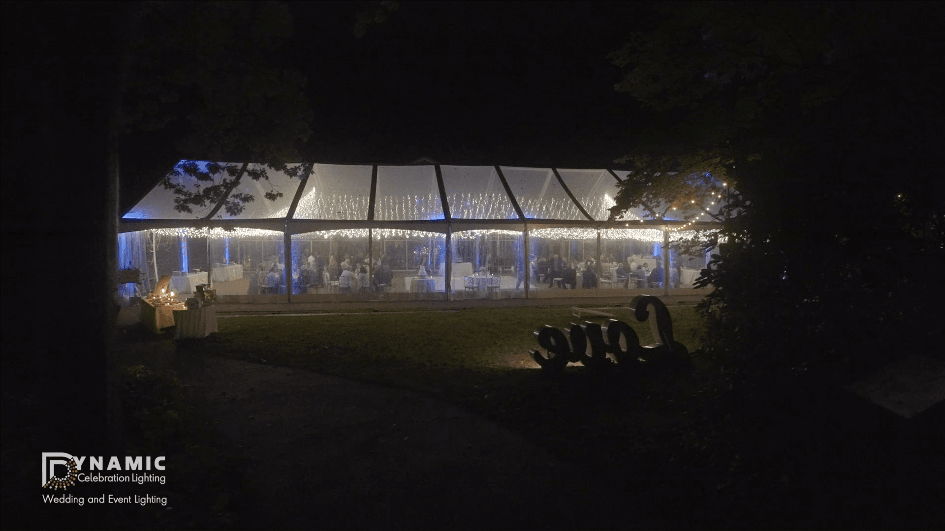 Large white event tent lit with string lights at night, seen from a distance. People are seated inside.