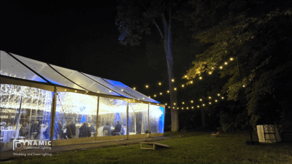 Outdoor wedding tent at night, lit with string lights, surrounded by trees and grass.