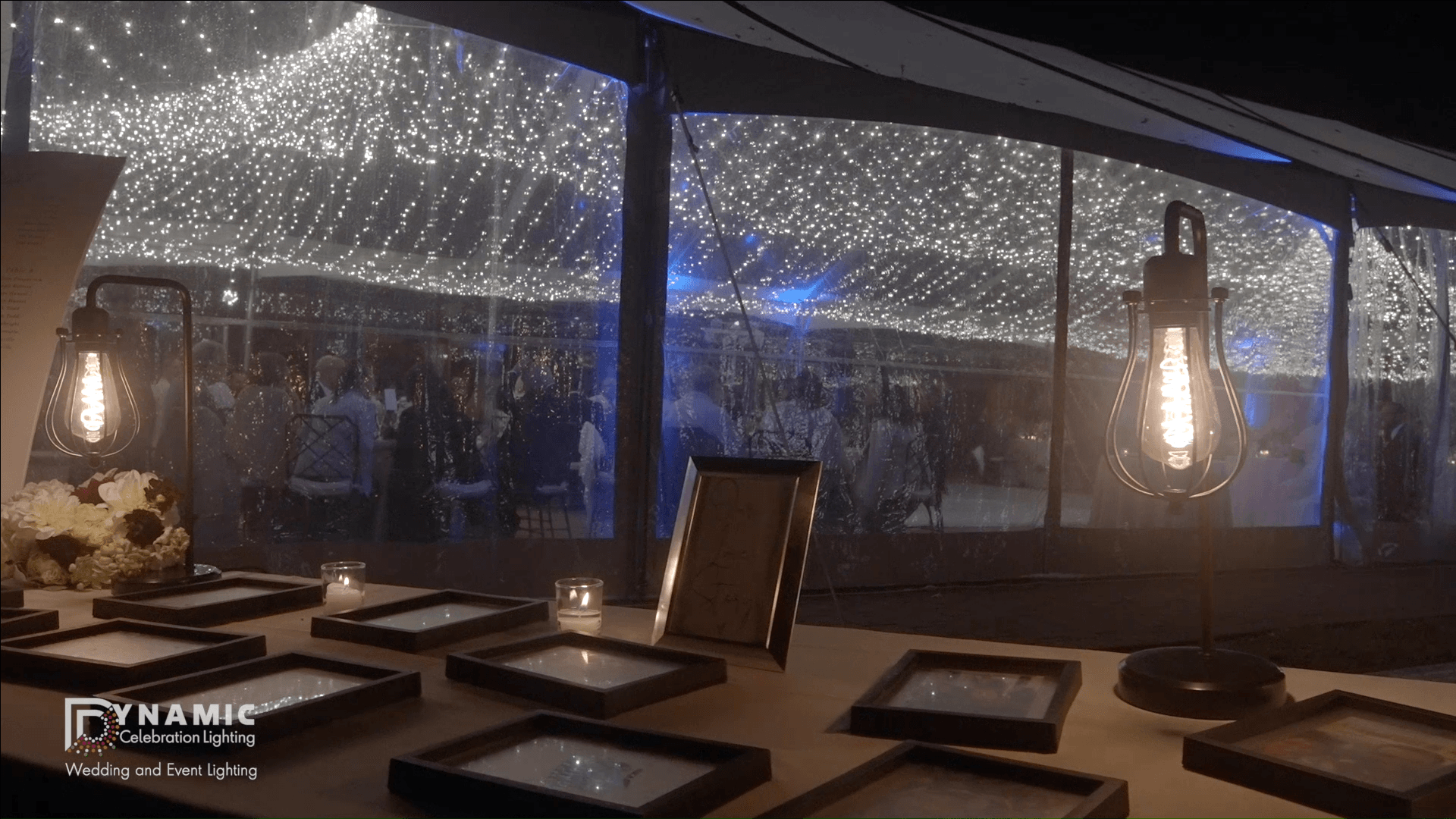 A warmly lit table with framed photos, candles, and string lights outside a tent with people inside at night.