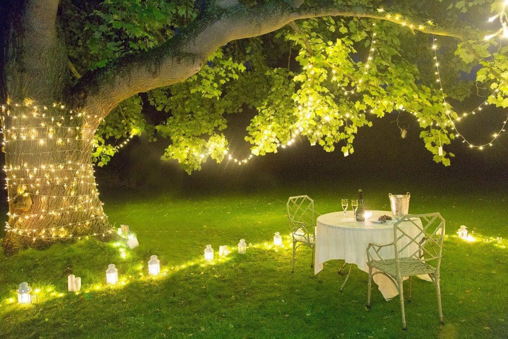 A round table for two under a tree with string lights, lanterns, and wine on a grassy lawn at night.