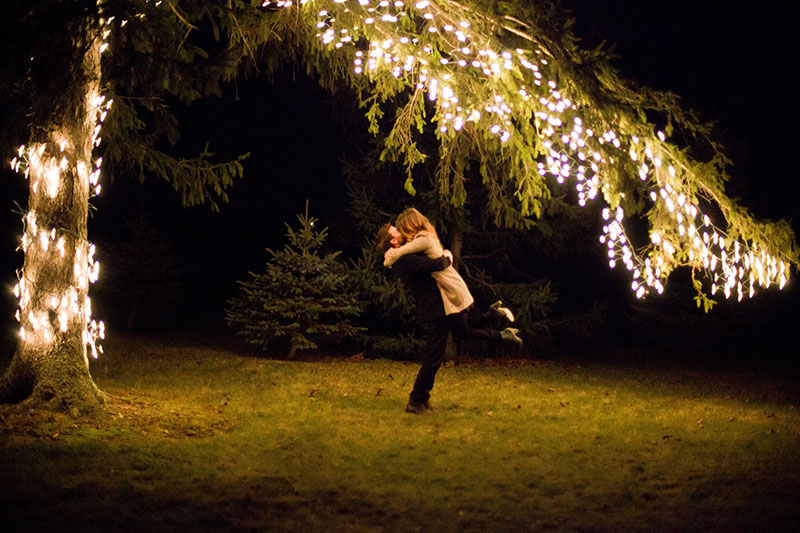 A couple embraces under a tree adorned with string lights at night, surrounded by darkness and grass.