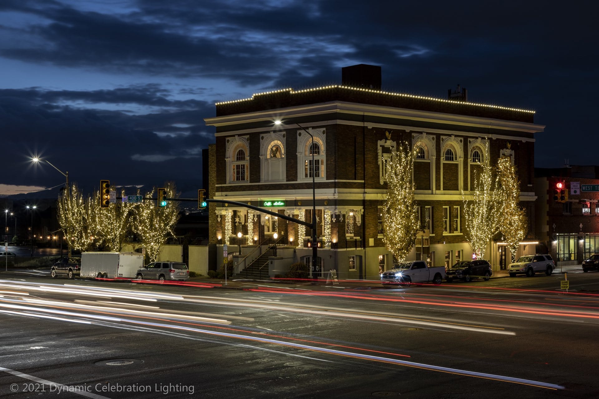 A brick building with trees wrapped in white holiday lights at dusk, with light streaks from passing cars.
